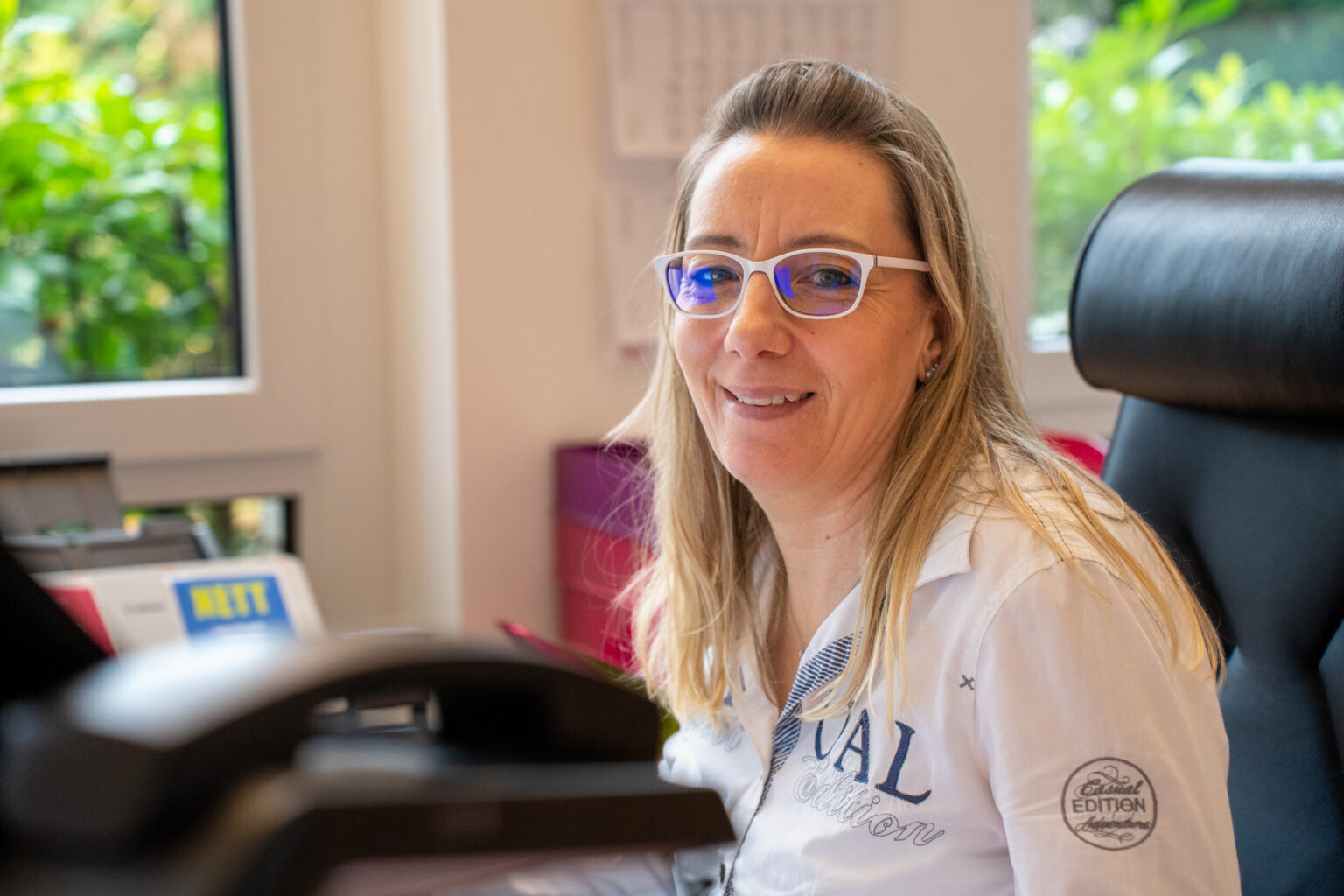 Frau mit Brille laechelt an ihrem Schreibtisch im Buero