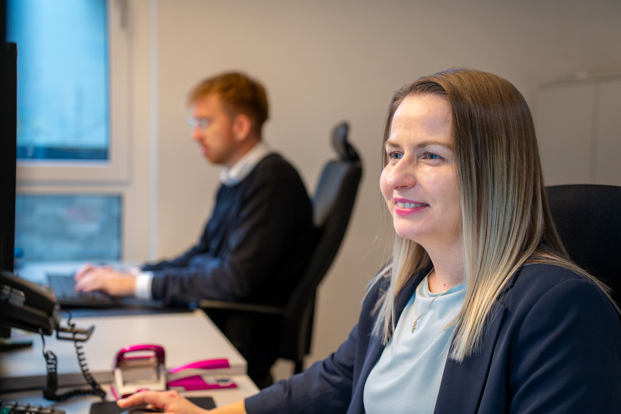 Frau sitzt an einem Schreibtisch, Mann arbeitet im Hintergrund am Computer