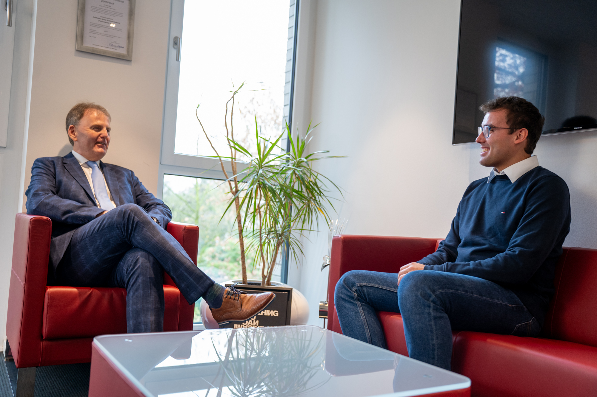 Zwei Männer sitzen in roten Sesseln und sprechen miteinander, Pflanze im Hintergrund